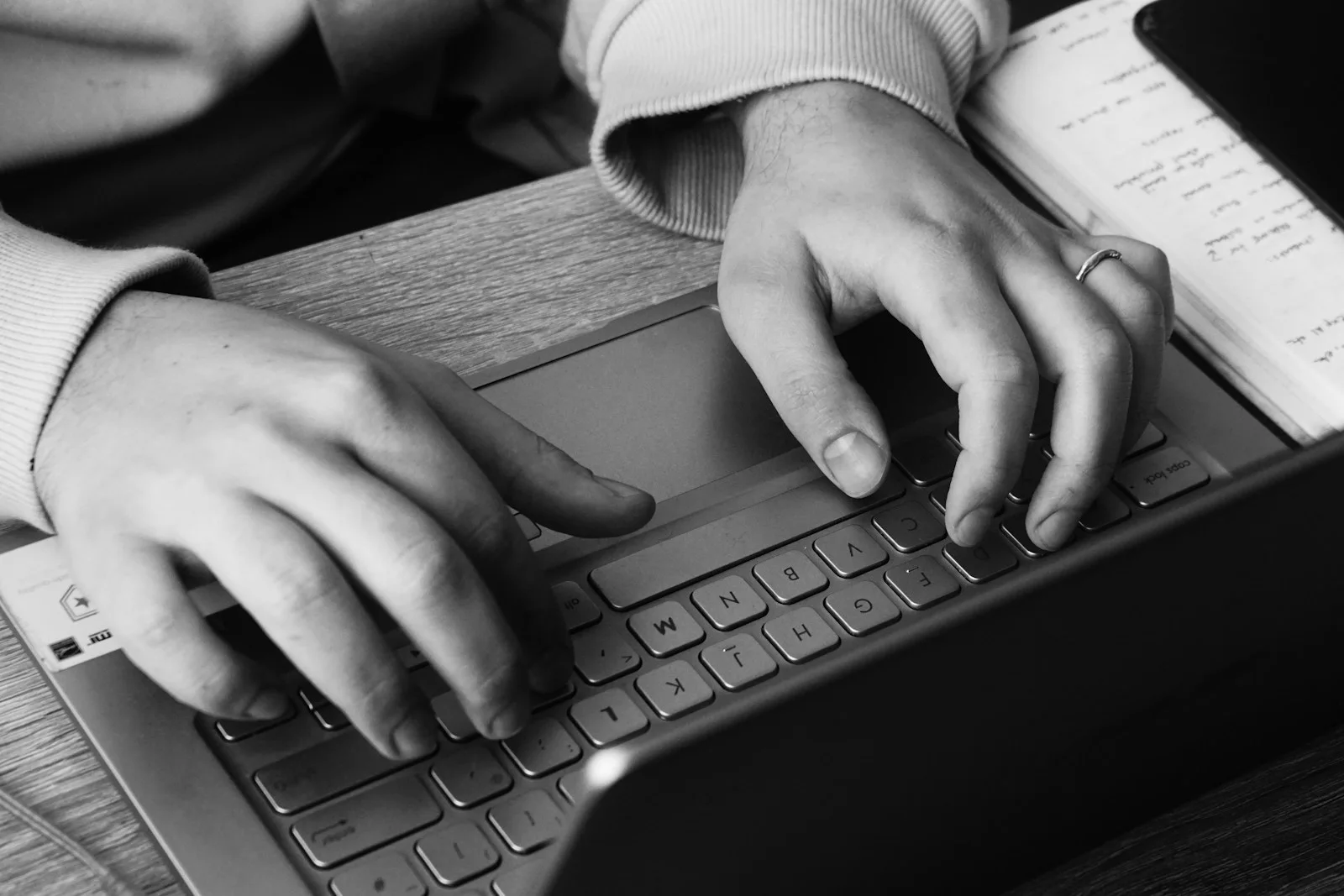 Black-and-white close-up of hands typing on a laptop beside handwritten notes