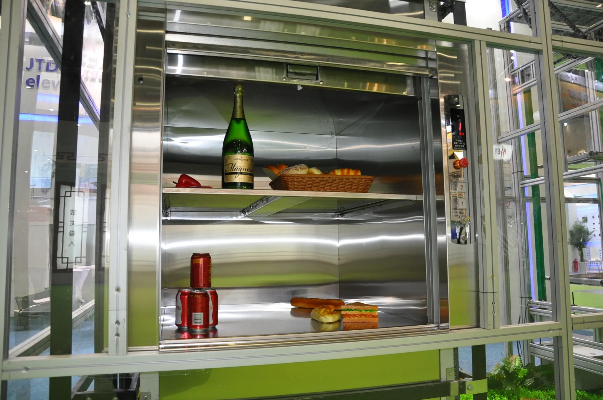 An open stainless-steel dumbwaiter car with shelves, display goods and landing controls.