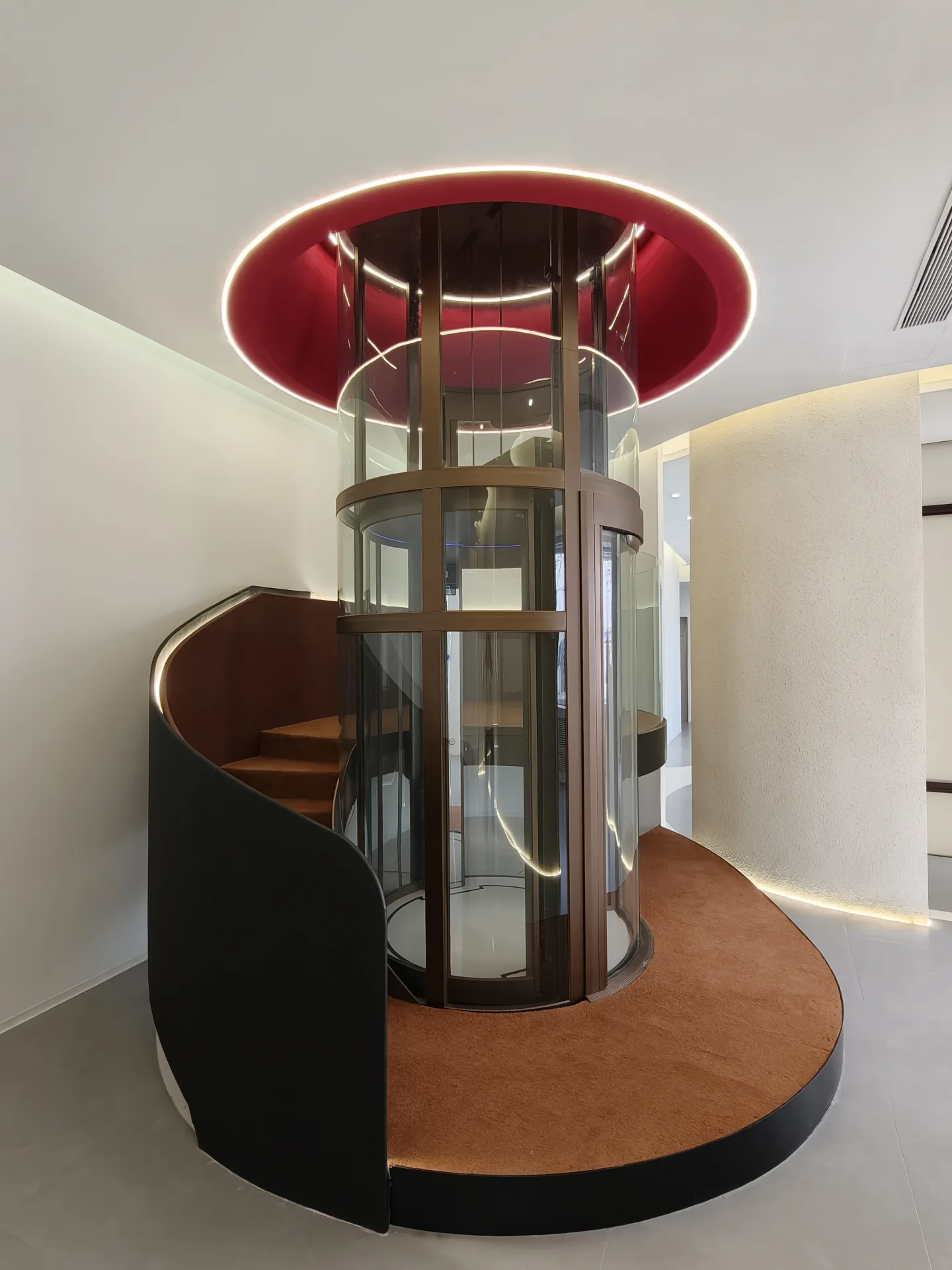 Front view of a round glass elevator beside a curved staircase in the showroom