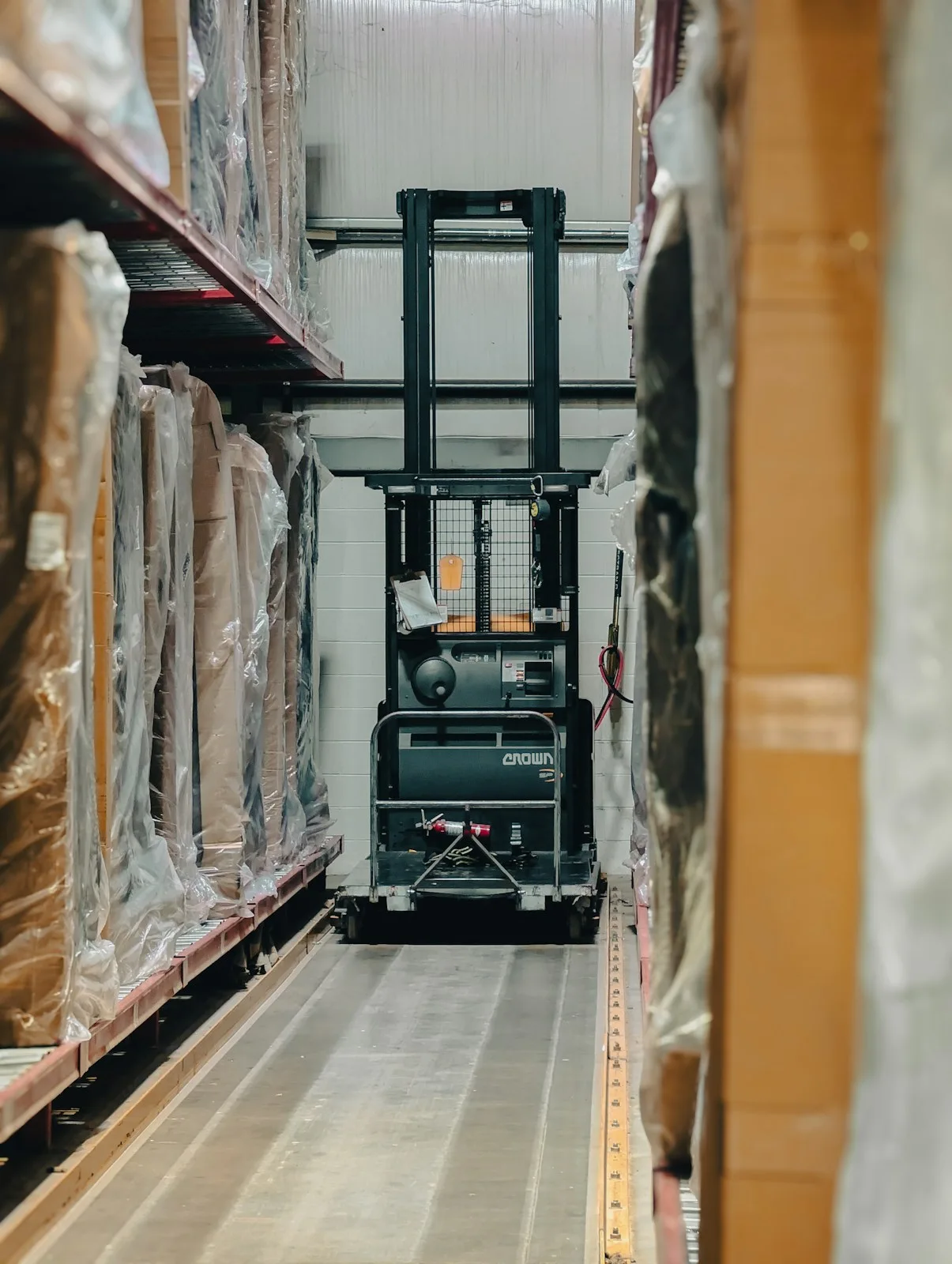 A forklift in a narrow warehouse aisle between shelves of stretch-wrapped pallets