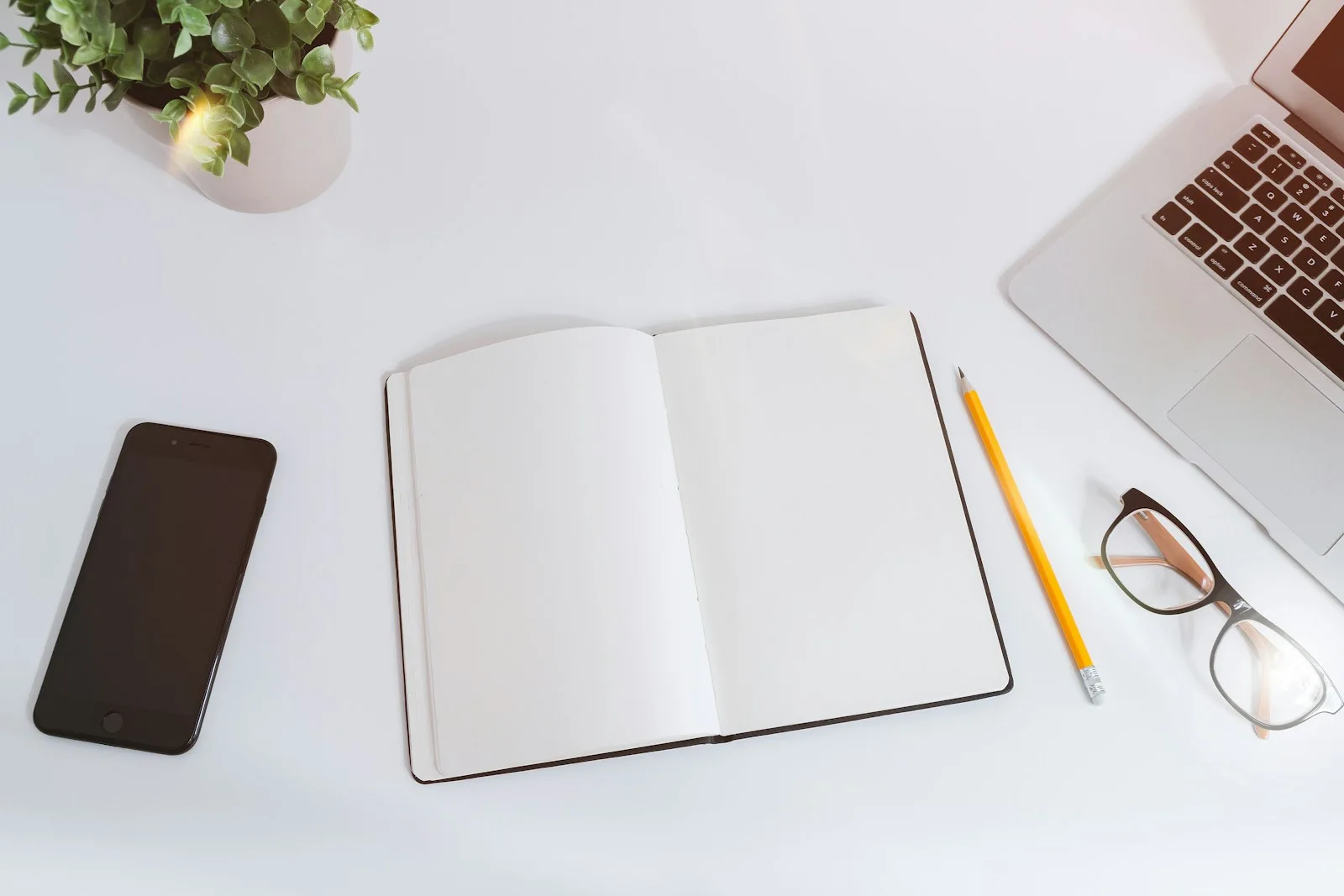 An open blank notebook with a pencil, phone, glasses and laptop on a white desk