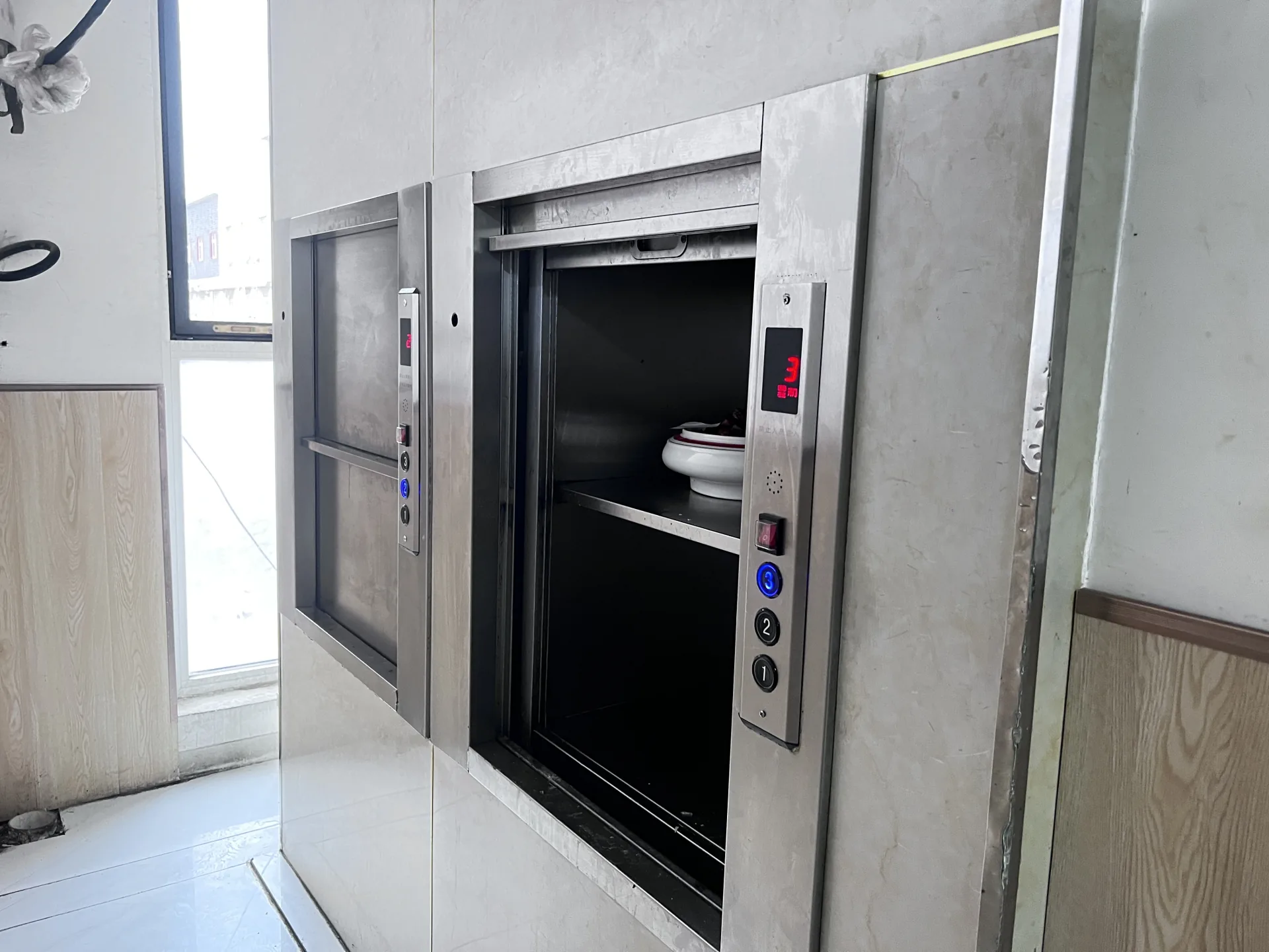 Counter-height stainless steel dumbwaiter with serving dishes on an open shelf